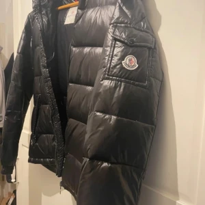 Svart Moncler pufferjacka med huva - Säljer en svart pufferjacka från Moncler med ikonisk logga på ärmen och coolt serietryck på insidan. Jackan har huva, stora fickor och grov dragkedja med Moncler-detalj. Perfekt för dig som vill ha en snygg och varm jacka med streetkänsla.