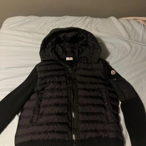 Svart dunväst med huva från Moncler - Säljer en svart dunväst från Moncler med huva och ribbstickade detaljer på sidor och rygg. Västen har dragkedja framtill, två fickor med knappstängning och Moncler-logga på vänster ärm. Materialet är en mix av dun och stickad polyester för extra komfort.  Har används bara 1 gång 
