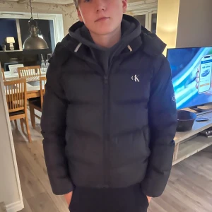Svart pufferjacka från Calvin Klein - Säljer en svart pufferjacka från Cal vin Klein med huva och dragkedja framtill. Jackan har CK-logga på bröstet och ärmen, samt en stilren, quiltad design. Perfekt för kalla dagar,   Jag köpte jackan ny på pondus förra året, för 2600kr