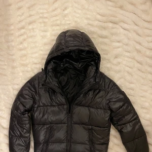 Lexington Svart pufferjacket med huva - Svart pufferjacka med stor huva och två dragkedjeförsedda fickor framtill. Jackan har quiltad design och är tillverkad i glansig syntet, perfekt för kalla dagar. Stängs med dragkedja och har en bra passform som ger en chill vibe. Värd 2500kr men säljes för 1000kr