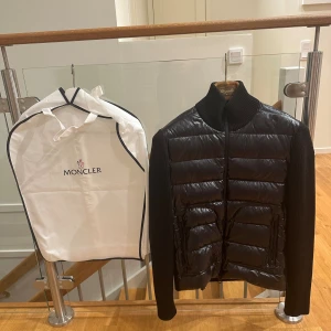 Moncler Tricot Cardigan Down Jacket Tricolor - Svart Cardigan från Moncler med quiltad front i blankt material och stickade ärmar samt rygg. Jackan har hög krage, två fickor med dragkedja och Moncler-logga på vänster ärm. Den är sparsamt använt och har inga skador, den är redan äkthetsverifierad av ”Vestitaire Collective” där den även är köptes ifrån. Lappar, dustbag och påse inkluderas.