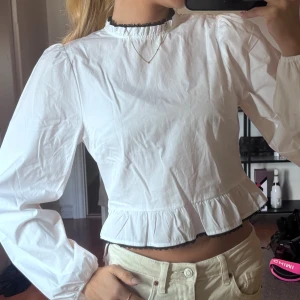 Vit blus med volang och svarta detaljer - Säljer en vit blus med lång ärm, volang nertill och svart spetskant vid hals och midja. Blusen har hög hals med knytband bak.
