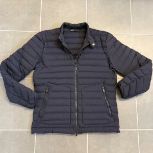 Mörkblå dunjacka från UBR, strl M - Snygg mörkblå dunjacka från UBR i storlek M. Jackan har ett stilrent, quiltat mönster, ståkrage med knappdetalj och två praktiska fickor med dragkedja. Perfekt för kyliga dagar och har en clean look som funkar till det mesta. Kvitto ingår!