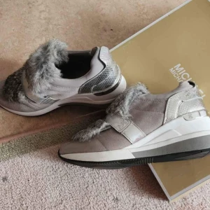 Michael Kors sneakers med pälsdetalj - Unika sneakers från Michael Kors i grå mocka med silvriga detaljer och fluffig grå päls över vristen. Skorna har en bred metallicrem och vit sula med svart undersida. Perfekt för dig som vill sticka ut med en lyxig och trendig look.