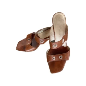 Bruna sandaler med blommor i skinn - Snygga bruna sandaler i skinn med fyrkantig tå och låg klack. Två breda remmar över foten dekorerade med vita broderade blommor ger en cool vintagekänsla. Perfekta för sommarens alla outfits och lätta att matcha med jeans eller klänning.