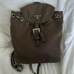  Prada ryggsäck  - Snygg brun ryggsäck från Prada i nylon med detaljer i skinn och silverfärgade metalldelar. Väskan har ett klassiskt trekantsmärke framtill, justerbara axelband i kedja och läder samt praktiskt innerfack med dragkedja. Obs väskan är inte stor. Granskad av experter hos vestiairecollective.com 