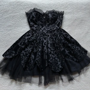 Svart kortklänning från Hell Bunny - Svart kortklänning med axelband, volangkant och dekorativ knapp framtill. Klänningen har ett mörkt, blommigt mönster och lager av tyll i kjolen för extra volym. Limited edition av märket Hell Bunny. Endast använd ett fåtal gånger 🖤