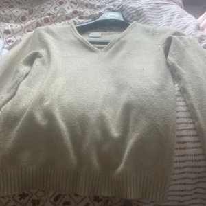 Beige v-ringad stickad tröja från Vila - Mysig beige stickad tröja från Vila med v-ringad hals och långa ärmar. Tröjan har ribbade muddar vid ärmslut och nederkant, vilket ger en snygg och enkel look. Perfekt för dig som gillar stilrena och neutrala färger.