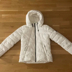 Vit pufferjacka med huva - Snygg vit pufferjacka från Save The Duck med svart dragkedja och huva med snörning. Jackan har två fickor framtill och ett svart logomärke på ärmen. Perfekt för kyliga dagar och enkel att matcha med olika outfits.