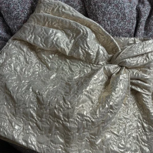 Champagnefärgad omlottkjol från Zara - Supersnygg champagnefärgad kortkjol från Zara med glansig, quiltad yta och omlottknytning framtill. Kjolen har en unik struktur och cool metallic-look som verkligen sticker ut. Perfekt för dig som vill ha något extra till outfiten.