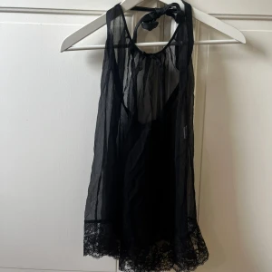 Svart transparent klänning linne topp/ nattlinne med spets - Säljer en svart, transparent nattlinne med knyt i nacken och vacker spetskant längst ner. Toppen är tunn och luftig med en lätt glansig finish, perfekt för att skapa en cool layering-look. Spetsdetaljen ger en extra touch av stil och edge. i 95% silkes 
