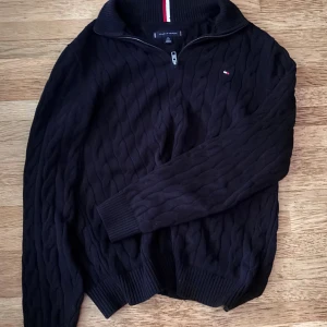 Svart stickad half zip från Tommy Hilfiger - Svart stickad tröja från Tommy Hilfiger med half zip och kabelstickat mönster. Liten logga på bröstet och ribbade muddar vid ärmslut och nederkant. Hög krage med Hilfigersignatur på insidan. Perfekt för dig som gillar klassisk stil.