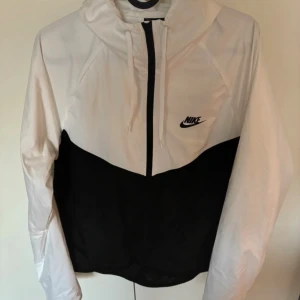 Nike svartvit vindjacka med huva - Nike vindjacka i svart och vitt med huva och dragkedja hela vägen. Jackan har två fickor med dragkedja och klassisk Nike-logga på bröstet. Tillverkad i lätt polyester, perfekt för blåsiga dagar. Snygg kontrast mellan svart och vitt, sportig och stilren look. Passar män och kvinnor därmed unisex, helt oanvänd. I storlek xl men kan passa runt 175cm.