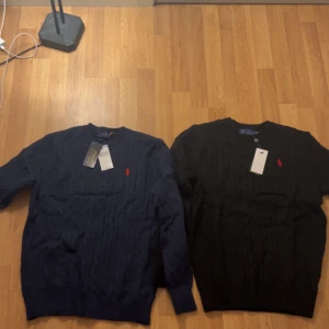 2 st svart,mörkblå polo Ralph Lauren tröjor - Svart kabelstickad tröja från Polo Ralph Lauren med klassisk rund hals och rött broderat logomärke på bröstet. Tröjan har lång ärm och ribbade muddar. Perfekt för dig som gillar stilren och tidlös design med premiumkänsla.