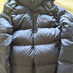 Mörkblå dunjacka från Polo Ralph Lauren - Snygg mörkblå dunjacka från Polo Ralph Lauren med röd broderad logga på bröstet. Jackan har hög krage, dragkedja framtill och flera praktiska fickor. Perfekt för kalla dagar och riktigt skön att bära. Klassisk och stilren design.