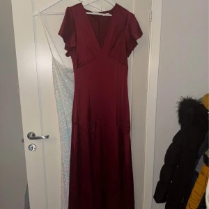 Vinröd långklänning från Bubbleroom - Säljer en vinröd långklänning från Bubbleroom med v-ringning och korta volangärmar. Klänningen har en elegant, flowy passform och är tillverkad i ett glansigt polyestertyg som ger en lyxig känsla. Perfekt för dig som vill sticka ut med en klassisk och stilren look.