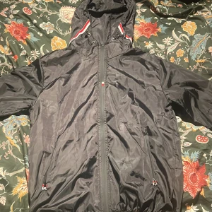 Svart vindjacka från Moncler med huva - Svart vindjacka från Moncler med huva och snygga rödvita detaljer på insidan av huvan. Jackan har dragkedja framtill, två sidofickor med dragkedja och Moncler-logga på ärmen. Materialet är lätt och syntetiskt, perfekt för blåsiga dagar.