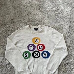 Vit stickad Stüssy tröja med färggrafik - Vit stickad tröja från Stüssy i storlek M med coolt tryck av biljardbollar i olika färger och bokstäver på bröstet. Rund hals och ribbade muddar vid ärmslut och nederkant. Perfekt för dig som gillar streetwear och vill sticka ut med grafiska detaljer.