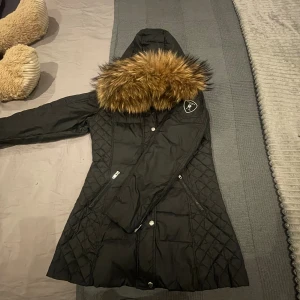 Svart Rockandblue dunjacka med päls - Snygg svart dunjacka från Rockandblue med avtagbar huva och fluffig brun päls. Jackan har quiltade detaljer på sidorna, dragkedjor framtill och ett Rockandblue-märke på ärmen. Perfekt för kalla dagar med sin långa, figurnära passform.