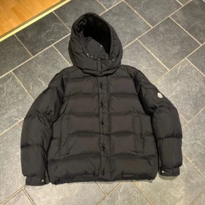 Svart Moncler dunjacka med huva - Säljer en svart dunjacka från Moncler med stor, justerbar huva och klassisk Moncler-logga på ärmen. Jackan har quiltad design, knappar och dragkedja framtill samt flera praktiska fickor. Perfekt för kalla dagar och riktigt snygg streetstil. Nyskick                   Storlek 3 motsvarar M/L