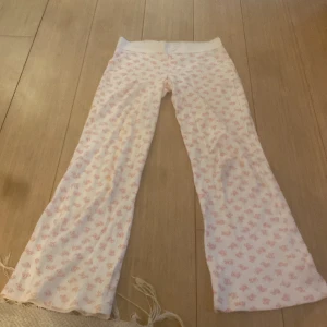 Vita pyjamasbyxor med rosa mönster - Mysiga vita pyjamasbyxor med ljusrosa mönster av små rosetter. Byxorna har en rak passform och resår i midjan för extra komfort. Perfekta för chill hemma eller sleepovers. Tillverkade i mjukt material som känns skönt mot huden.