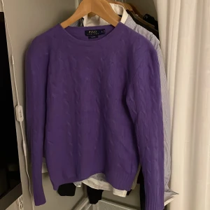Lila kashmirtröja från Polo Ralph Lauren - Supermjuk lila kabelstickad tröja från Polo Ralph Lauren i lyxig kashmir. Klassisk rund halsringning och långärmad modell, perfekt för att hålla värmen under kyliga dagar..