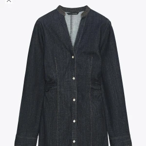 Mörkblå jeansklänning med knappar - Snygg mörkblå jeansklänning från Zara med långa ärmar och markerad midja. Klänningen har v-ringning, synliga kontrastsömmar och knappar hela vägen framtill. Perfekt för dig som gillar stilren denim med en modern twist. Prislapp kvar helt ny. 