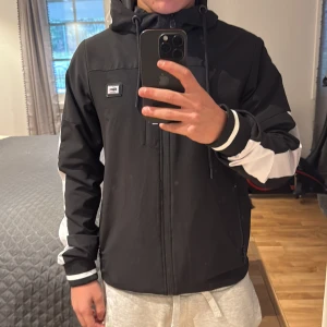 Svart och vit vindjacka från Lacoste - Snygg svart och vit vindjacka från Lacoste med huva och dragkedja framtill. Jackan har coola vita detaljer på ärmar och huva, samt Lacoste-logga på bröstet. Perfekt för dig som gillar sportig och stilren look. Meshfoder på insidan för extra komfort.