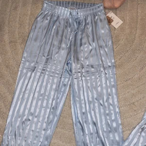 Blå randiga satinbyxor med snörning - Snygga blå satinbyxor med glansig finish och vertikala ränder. Byxorna har elastisk midja med snörning för skön passform och raka ben. Perfekta för chill hemma eller som pyjamas. Mjuk känsla och stilren look.