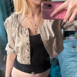 Beige bolero från Zoul - Säljer en beige bolero från Zoul med rundad hals och puffiga långärmar. Boleron har knappar framtill och resår vid midjan och ärmarna för en snygg passform. Materialet är glansigt och känns lätt mot huden, perfekt för att lyfta din outfit.