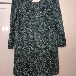 Klänning - Snygg klänning från Jacqueline de Yong i storlek 38. Klänningen har ett mörkt, abstrakt mönster i blått och grönt, v-ringad hals och långa ärmar. Tillverkad i mjuk polyester som känns skön mot huden. Perfekt för dig som gillar unika prints och bekväm passform.