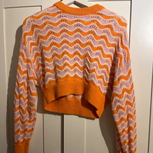 Croppad stickad tröja i orange och rosa - Superfin croppad stickad tröja med vågigt mönster i orange och ljusrosa. Tröjan har lång ärm, ribbade muddar och en loose fit. Perfekt för dig som vill sticka ut med färg och mönster!