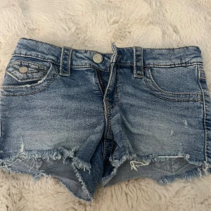Blå jeansshorts från Gina Tricot - Snygga blå jeansshorts från Gina Tricot med fransig kant och slitna detaljer. Klassisk femficksmodell med bälteshällor och knappstängning. 