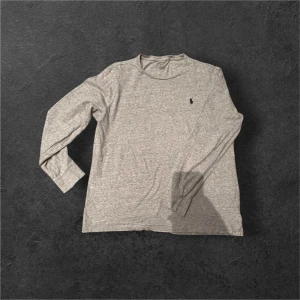 Grå långärmad tröja från Polo Ralph Lauren - Säljer en stilren grå långärmad t shirt från Polo Ralph Lauren med klassisk rund hals och den ikoniska svarta loggan broderad på bröstet. Tröjan är i mjuk bomull och passar perfekt till jeans eller chinos.