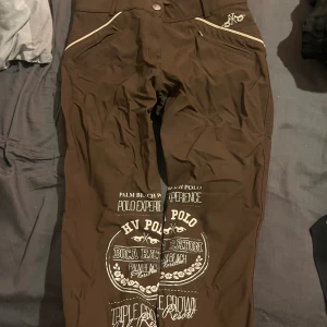 Bruna HV Polo täckbyxor med tryck ridbyxor  - Snygga bruna täckbyxor från HV Polo med vita kontrastdetaljer och stora tryck på framsidan. Byxorna har fickor med vit piping och ett lädermärke bak i midjan. Perfekta för dig som gillar sportig stil och vill sticka ut med coola prints.