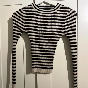 Randig ribbad tröja i svart och beige - Säljer en tight ribbstickad tröja med svarta och beiga ränder. Tröjan har lång ärm, rund halsringning och är stretchig för en snygg passform. Perfekt att styla med jeans eller kjol för en trendig look.