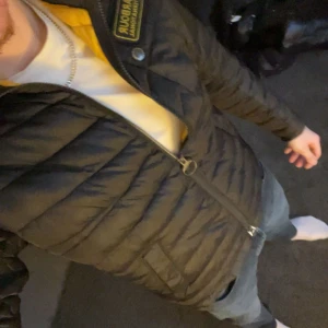 Svart pufferjacka Barbour herr - Svart pufferjacka från Barbour med quiltad design och ståkrage. Jackan har en snygg Barbour-logga på bröstet, tvåvägsdragkedja och långärmad passform. Perfekt för dig som gillar stilren och sportig look under höst och vinter.