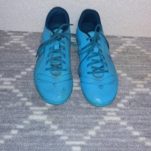 Blå Nike fotbollsskor med dobbar - Säljer ett par blå Nike fotbollsskor med snörning och orangea dobbar. Skorna har en sportig design med Nike-logga på sidorna och är tillverkade i syntetmaterial. Perfekta för dig som vill ha snygga och funktionella skor på planen.
