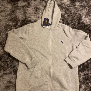 Grå kofta från Polo Ralph Lauren - Snygg grå hoodie från Polo Ralph Lauren med dragkedja och huva. Tröjan har lång ärm, två fickor fram och den klassiska Polo-loggan broderad på bröstet. Slim fit-modell i mjuk bomullsmix som är skön att bära. Perfekt för chill dagar.