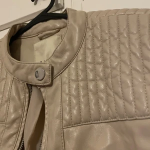 Beige skinnjacka från Amisu - Snygg beige skinnjacka från Amisu med quiltade detaljer på axlarna och dragkedjor vid fickor och ärmslut. Jackan har en rund krage med knapp och är helfodrad. Perfekt för dig som gillar stilrena och coola jackor.