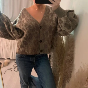 Beige och brun kofta med knappar - Säljer en mysig beige och brun kofta med v-ringad hals och stora knappar framtill. Koftan har ett mönster som påminner om leopard och är croppad med breda ribbade muddar vid ärmslut. Perfekt för dig som gillar chill och trendiga plagg.