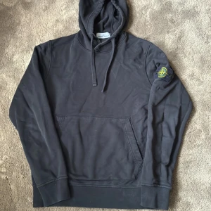 Svart hoodie från Stone Island - Svart hoodie från Stone Island med klassisk patch på vänster ärm. Tröjan har huva med snörning och en stor magficka framtill.