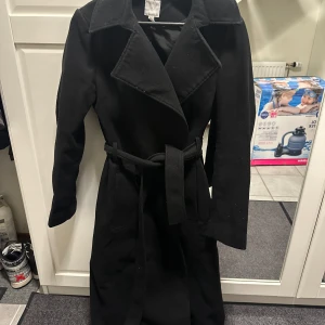 Svart kappa med bälte från H&M - Säljer en svart lång kappa från H&M i storlek 34. Den har klassisk krage, långa ärmar och ett snyggt bälte i midjan.