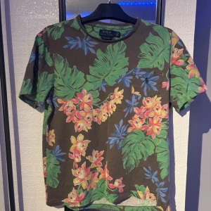 Blommig t-shirt från Polo Ralph Lauren - Snygg brun t-shirt från Polo Ralph Lauren med färgglatt blommönster i grönt, blått, rosa och gult. Modellen är custom slim fit och har en bröstficka. Perfekt för dig som vill sticka ut med en tropisk vibe.