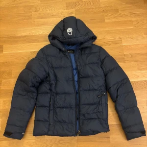Mörkblå pufferjacka från Sail Racing - Säljer en mörkblå pufferjacka från Sail Racing i storlek XS. Jackan har huva med snörning, dragkedja framtill och två sidofickor med dragkedja. Skulle säga att den är lite större än Xs och lite mer i S (stor i storlek) Väldigt varm och skön under vintern. Inget nyskick och därav priset.