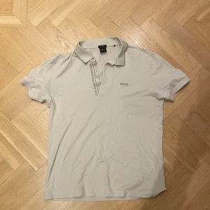 Beige slim fit pikétröja från BOSS - Snygg beige pikétröja från BOSS i slim fit med klassisk krage och korta ärmar. Tröjan har BOSS-logga broderad på bröstet samt tryckt längs knappslån. Perfekt för dig som gillar stilrena och enkla plagg. Använd men i bra skick (behöver dock strykas:)).