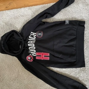 Svart hoodie från Hoodrich XS - Svart hoodie från Hoodrich i storlek XS med stora vita och röda tryck och broderade märken på bröstet. Tröjan har huva med snörning, känguruficka och lång ärm. Materialet är mjukt och känns som bomull med fleece på insidan. Snyggt Hoodrich-tryck även på ärmen.