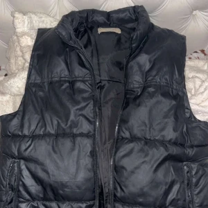 Svart dunväst med dragkedja - En svart, quiltad dunväst med hög krage och dragkedja framtill. Västen har två fickor med dragkedja på sidorna och en stilren, enkel design. Perfekt för lager-på-lager och passar till många olika outfits.