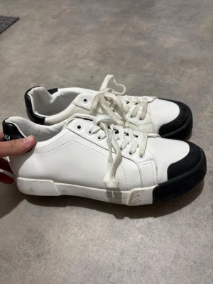 Vita Dolce & Gabbana sneakers - Snygga vita sneakers från Dolce & Gabbana med svarta detaljer på tå och häl. Skorna har klassisk låg profil, vita snören och är tillverkade i slätt läder. Perfekta för dig som vill ha en clean och stilren look med exklusiv känsla.