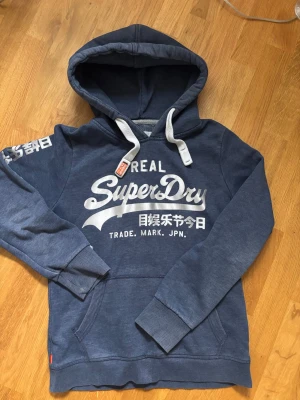 Blå Superdry hoodie med tryck - Personlig favorit från droppet.               Snygg blå hoodie från Superdry med stort vitt tryck på bröstet och japanska tecken på ärmen. Hoodien har en klassisk huva med vita dragsnören och ribbade muddar vid ärmar och nederkant. Perfekt för en avslappnad och trendig stil.